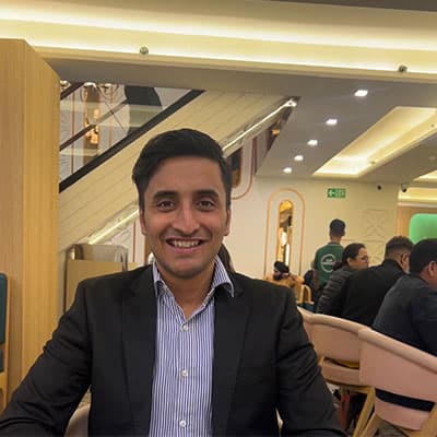 A headshot of Ayush Garg wearing a suit and shirt. He is smiling at the camera.